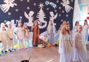 Dzieci w strojach jasełkowych stoją w półkolu, a na środku dwie dziewczynki. Przedszkolaki śpiewają piosenkę "Najpiękniejsze dni w roku".