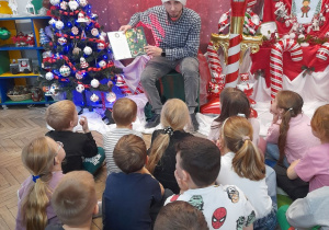 Dzieci siedzą na dywanie, a Pan Łukasz prezentuje dzieciom obrazek w książce. W tle dekoracja świąteczna