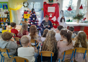 Dzieci siedzą na krzesełkach, a przed nimi Pan Mateusz czyta opowieść pt. "Wolny dzień Mikołaja" z książki "Opowiadania na Boże Narodzenie" tłumacząc niektóre słowa na język angielski.