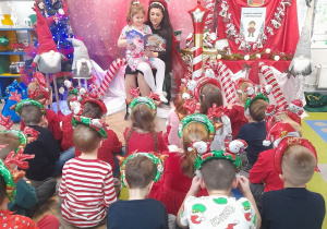 Dzieci siedzą na dywanie w świątecznych opaskach, a mama Nadii czyta dzieciom książkę pt. "Idą święta". W tle dekoracja mikołajkowa.