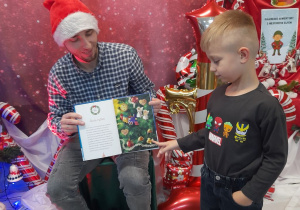 Pan Łukasz siedzi na krześle i trzyma książkę. Obok stoi Wiktor i pokazuje na obrazku tytułową złotą rybkę.