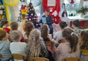 Dzieci siedzą na krzesełkach, a przed nimi Pan Mateusz - nauczyciel języka angielskiego, który prezentuje książkę pt. "Opowiadania na Boże Narodzenie".