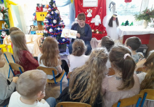 Pan Mateusz siedzi obok choinki i pokazuje dzieciom obrazek w książce.