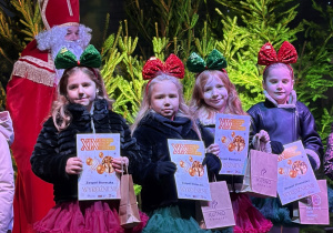 Zespół "Słoneczka" stoi na scenie z dyplomami i upominkami. Obok stoi Mikołaj. W tle dekoracja świąteczna.