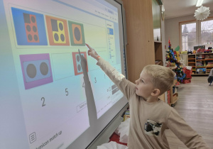 Lucjan stoi przy tablicy multimedialnej. Chłopiec dopasowuje kształt klocka Numicon do odpowiedniej cyfry.