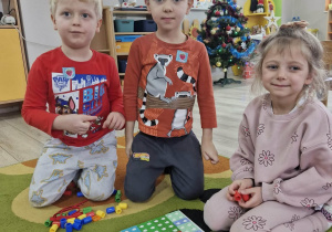 Karol, Wojtek i Liliana siedzą na dywanie. Przed dziećmi leżą ułożone na planszy klocki Numicon.