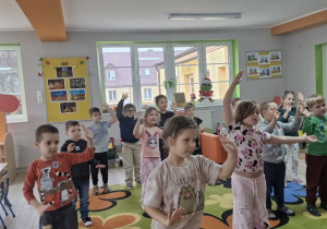 Dzieci stoją na dywanie z rączkami w górze w trakcie zabawy ruchowej.