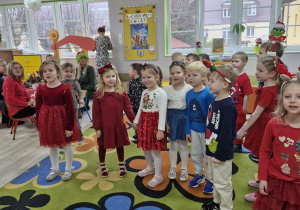 Przedszkolaki w odświętnych strojach stoją na dywanie i śpiewają kolędę.