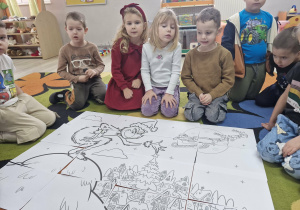 Na dywanie leży bardzo duża kolorowanka z wizerunkiem Grincha. Wokół siedzą przedszkolaki.