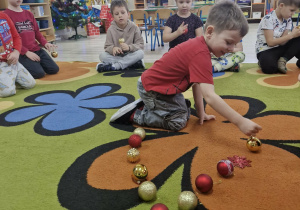 Dzieci siedzą na dywanie. Antoś układa bombki w kształt trójkąta na dywanie.