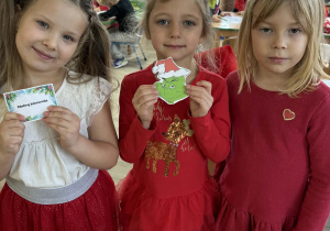 Maja, Lila i Nadia w świątecznych strojach pozują do zdjęcia. Dziewczynki mają w dłoniach kartkę z zadaniem od Grincha i obrazek Grincha.