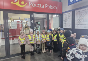Dzieci w odblaskowych kamizelkach stoją przed budynkiem Poczty Polskiej.