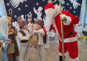 Mikołaj wręcza upominki dzieciom. W tle jasełkowa dekoracja.