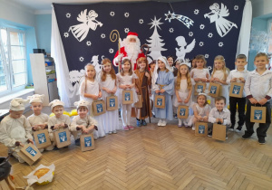 Dzieci w strojach jasełkowych pozują do zdjęcia z Mikołajem. W tle jasełkowa dekoracja.