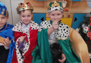 Trzech chłopców w roli Królów siedzi obok siebie. Dzieci trzymają kadzidło, złoto i mirrę.