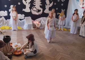 Tosia w roli Aniołka stoi na środku sceny. W tle fragment dekoracji jasełkowej, dziewczynki w roli Aniołków i Gwiazdka. Z prawej strony Mikołaj w roli Gospodarza, Król, z lewej siedzą Baranki i Pastuszkowie przy ognisku.