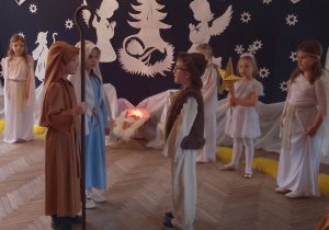 Marysia w roli Maryi i Antek w roli Józefa stoją przed Mikołajem w roli Gospodarza. W tle fragment dekoracji jasełkowej i dziewczynki w roli Aniołków oraz Gwiazdki