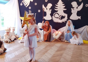 Zuzia w roli Gwiazdki stoi na środku sceny. W tle Józef z Maryją siedzą przy żłóbku, a obok śpią Aniołki. Z lewej strony siedzą Baranki i Pastuszek.