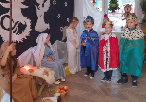 Królowie stoją obok żłóbka z darami w rękach. Przy żłobku siedzą Maryja i Józef, a obok stoi Aniołek. W tle dekoracja jasełkowa.