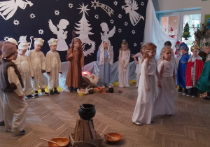 Dzieci w strojach jasełkowych stoją w półkolu, a na środku dwie dziewczynki. Przedszkolaki śpiewają piosenkę "Najpiękniejsze dni w roku".