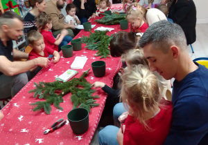 Rodzice i dzieci siedzą przy stoliku przykrytym czerwonym obrusem. Na stole jodłowe gałązki, doniczki i sekatory.