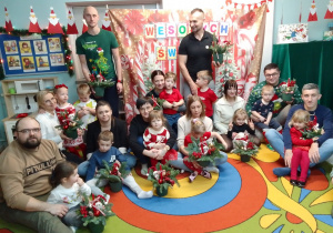 Rodzice i dzieci z gotowymi stroikami pozują do wspólnego zdjęcia na tle świątecznej dekoracji.