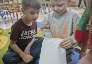 Dzieci siedząc na dywanie otwierają kopertę z Grinchem.