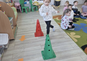 Zuzia porusza się po torze przeszkód. Na podłodze widać pachołki i ułożone pomarańczowe strzałki.