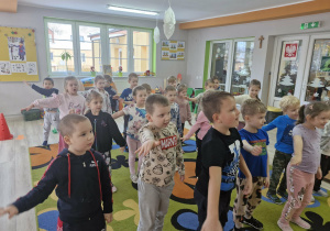 Dzieci stoją na dywanie w trakcie zabawy ruchowej i wyciągają na bok prawe dłonie.