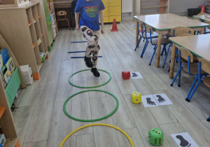 Oluś skacze na jednej nodze w jednej z dużych obręczy leżących na podłodze. Obok widać kostki do gry i obrazki z wizerunkami stóp.