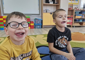 Antoś i Staś siedząc na dywanie wysuwają język na boki.