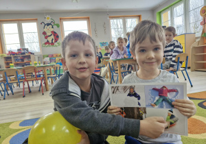 Ignacy trzyma w dłoni planszę z obrazkami, Franciszek wskazuje na fragment przedstawiający jazdę na sankach.