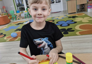 Oluś siedzi przy stoliku. Chłopiec mazakiem ozdabia wykonane z kolorowego papiery łyżwy.