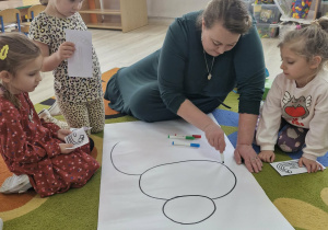 Pani z Biblioteki Pedagogicznej siedzi z dziećmi na dywanie i rysuje mazakami na dużym białym kartonie.