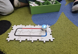 Na dywanie widać ułożone puzzle z wizerunkiem drogi i jeżdżącego na nich ozobota.