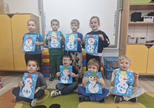 Dzieci stoją przed tablicą multimedialną i prezentują wykonane prace plastyczne - bałwanki wykonane z papieru kolorowego i gotowych elementów.