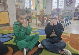 Staś i Antoś siedzą na dywanie. Chłopcy przykładają obie dłonie do ust.