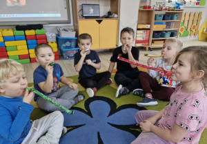 Dzieci siedzą na dywanie i dmuchają w gwizdki urodzinowe.