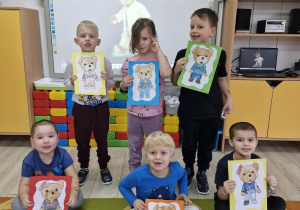 Przedszkolaki prezentują wykonane prace plastyczne. W tle na tablicy multimedialnej widać postać Misia Uszatka.