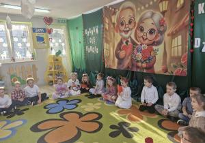 Dzieci siedzą na dywanie czekając na rozpoczęcie występu. W tle widać dekorację przygotowaną z okazji Dnia Babci i Dziadka.