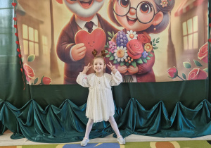 Zuzanna pozuje do zdjęcia na tle dekoracji przygotowanej z okazji Dnia Babci i Dziadka.