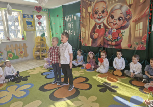 Lucjan i Franciszek stoją na środku sceny aby powitać przybyłych gości. Za chłopcami na dywanie siedzą pozostałe dzieci.