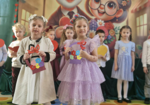 Liliana i Klara składają życzenia zebranym babciom i dziadkom. Dziewczynki trzymają w dłoniach papierowe koszyczki z kwiatkami.