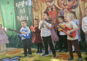 Szymon, Franek i Karol stoją na środku sceny. Chłopcy trzymają w dłoniach instrumenty muzyczne (gitary i trąbkę), na których grają koncert dla swoich dziadków.