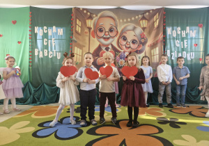 Zuzia, Szymon, Karolek i Alicja stoją na środku sceny. Dzieci trzymają w dłoniach czerwone papierowe serduszka.