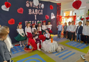 Mikołaj w roli Dziadka siedzi na kanapie odwrócony do Nadii w roli Babci. Chłopiec śpiewa fragment piosenki. W tle rówieśnicy stoją na tle dekoracji.