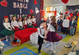 Dzieci stoją w półkolu na tle dekoracji i śpiewają piosenkę, a Nadia w roli Babci tańczy z Mikołajem w roli Dziadka w kółeczku.