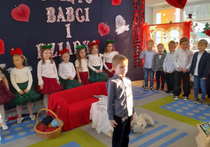 Adaś stoi na środku sceny i zaprasza Babcie i Dziadków na występ. W tle koleżanki i koledzy z grupy stoją na tle dekoracji.