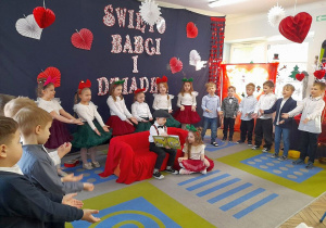 Na kanapie siedzi Mikołaj w roli Dziadka. Obok kanapy siedzi Amanda w roli wnuczki. Dziadek pokazuje wnuczce książkę, a pozostałe dzieci śpiewają piosenkę "Dziadkowe bajeczki". W tle dekoracja.