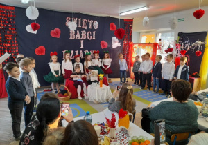 Dzieci stoją w półkolu na tle dekoracji i śpiewają piosenkę. Nadia w roli Babci siedzi na kanapie i robi na drutach. Obok Mikołaj w roli Dziadka czyta gazetę.